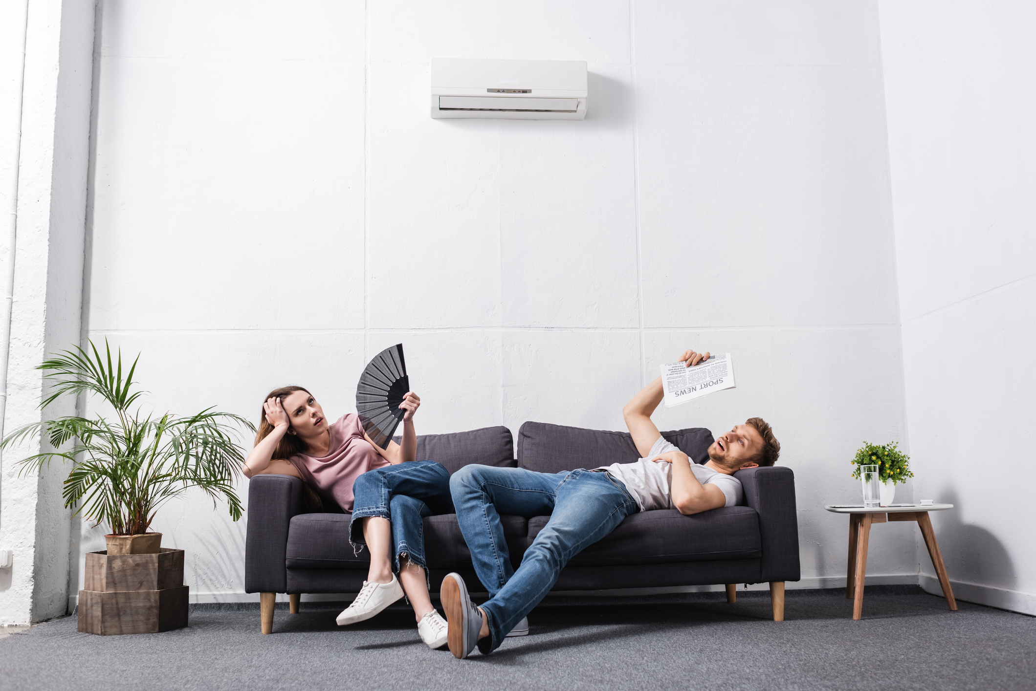 young couple with hand fan and newspaper suffering from heat at home with broken air conditioner young couple with hand fan and newspaper suffering from heat at home with broken air conditioner